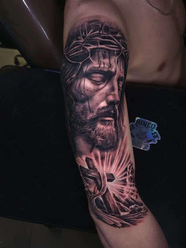 Tatuagem de Jesus Cristo com coroa de espinhos e cruz em realismo no braço