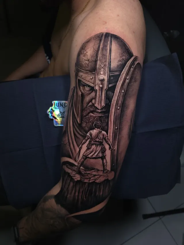 Tatuagem de guerreiro com elmo e espada em realismo preto e cinza no braço